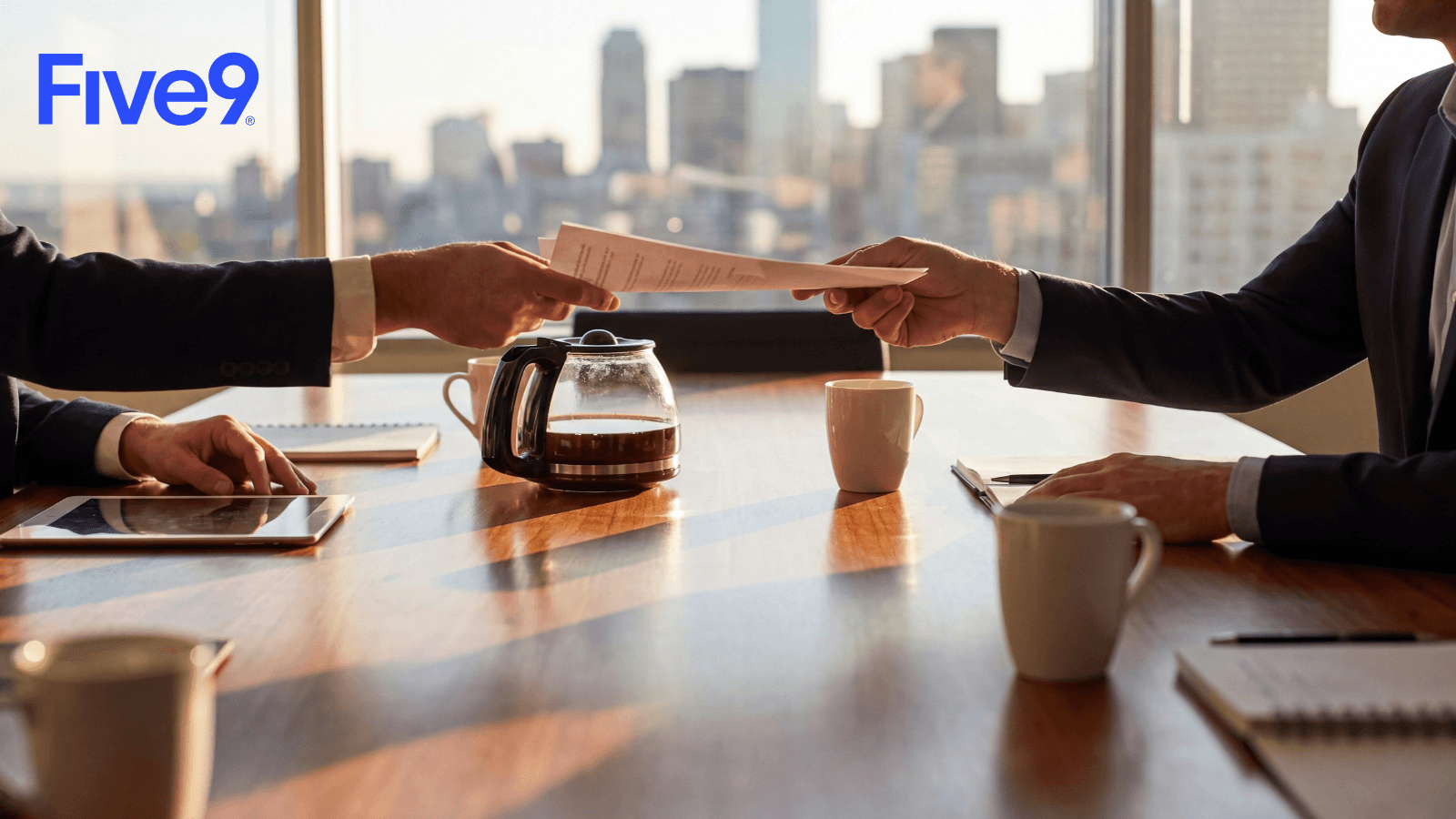 Two businesspeople pass a document across a boardroom table with coffee cups and a city skyline behind them, with the Five9 logo in the corner