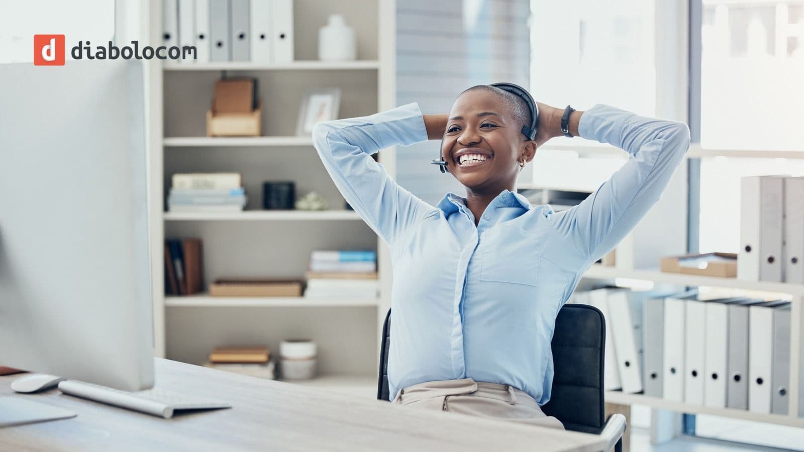 Smiling contact center agent wearing a headset leans back in her chair with her hands behind her head at a tidy desk in a bright office, with the Diabolocom logo in the top left