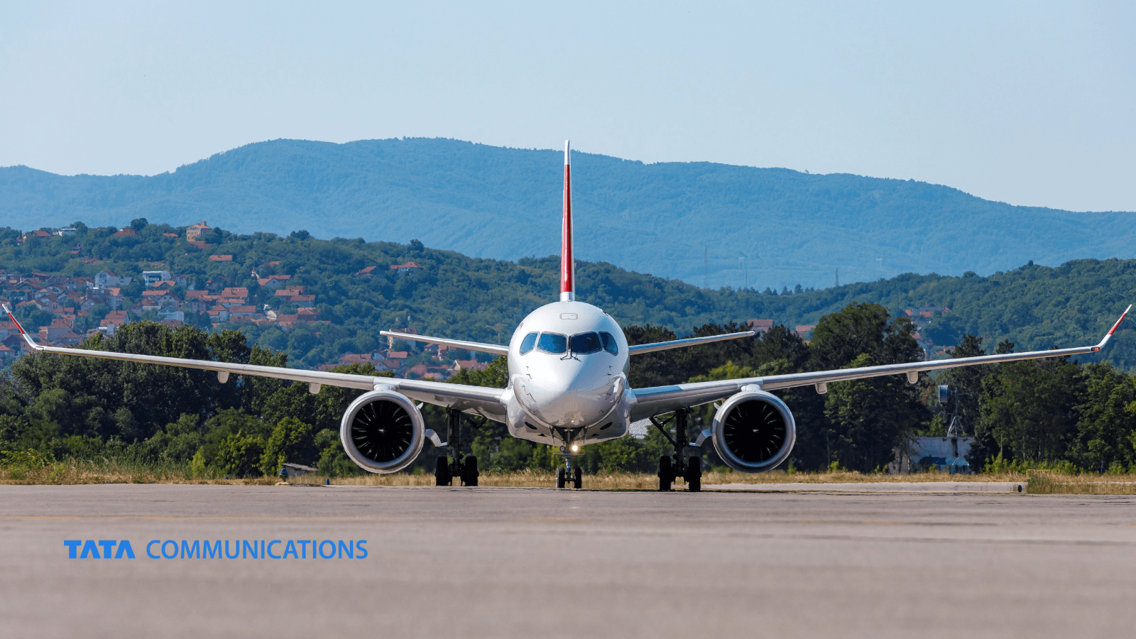 A passenger jet faces the camera on a runway ahead of take-off, with green hills behind and the Tata Communications logo in the lower left.