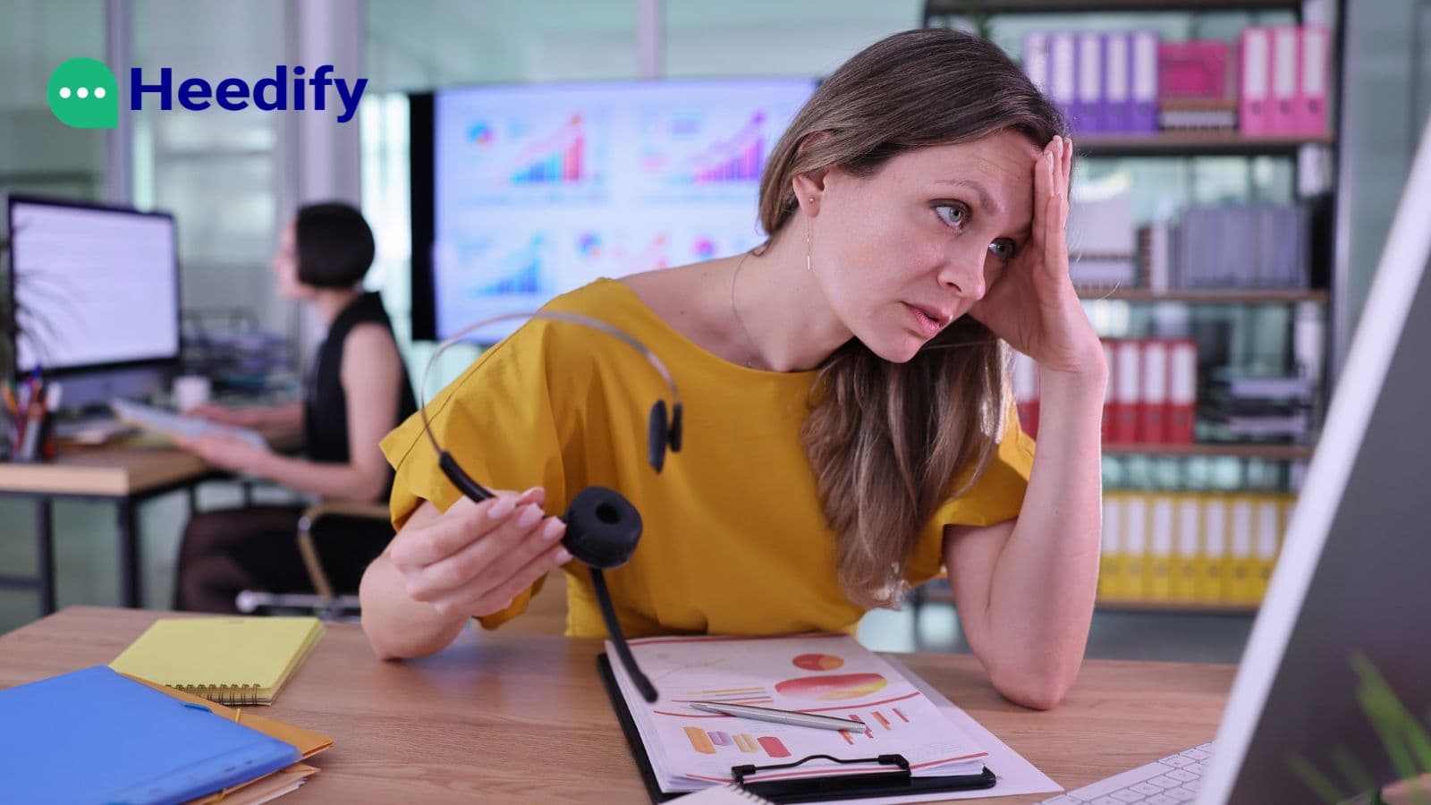 A frustrated contact centre agent in a yellow top holds her headset away from her ear and rests her head on her hand at her desk. A colleague works at a computer behind her. The Heedify logo appears in the top left corner