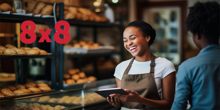 A young female employee greets customers with a warm smile as they enter the bustling bakery, its shelves brimming with the aroma of freshly baked bread and pastries.